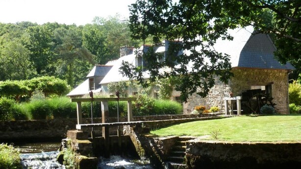 A rustic stone building with a small bridge over a stream, surrounded by lush greenery.