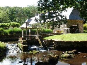 A rustic stone building with a small bridge over a stream, surrounded by lush greenery.