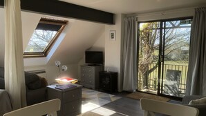 Un salon avec un plafond en pente, un téléviseur posé sur un meuble, une table recouverte d’une nappe et une vue sur l’extérieur grâce à de grandes fenêtres.