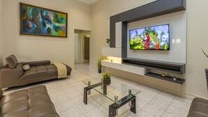 A modern living room with a large flat-screen TV mounted on the wall, a brown leather sofa, a glass coffee table, and a colorful abstract painting. - Sleeps 16 @ Palm Beach - 6BR Villa w/ Private Pool (Palm Beach)