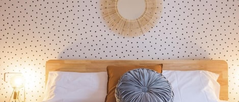 A neatly made bed with white linens, a wooden headboard, and two pillows, one brown and one gray, in a room with a polka dot wall and a circular mirror above the bed.