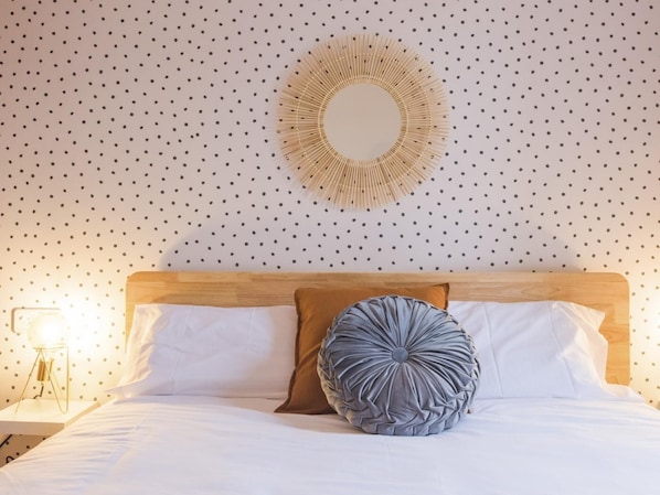 A neatly made bed with white linens, a wooden headboard, and two pillows, one brown and one gray, in a room with a polka dot wall and a circular mirror above the bed. - La Ventana de Leonor (Toledo)