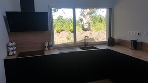 A modern kitchen with a built-in stove, a sink, and a window overlooking a garden. - Maison Neuve Proche mer Avec Piscine Chauffée (Fouesnant)
