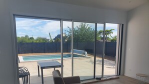 A living room with a large sliding glass door leading to a backyard with a swimming pool. - Maison Neuve Proche mer Avec Piscine Chauffée (Fouesnant)