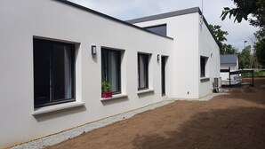 A modern building with white exterior walls, multiple windows, and a small garden area with a potted plant. - Maison Neuve Proche mer Avec Piscine Chauffée (Fouesnant)