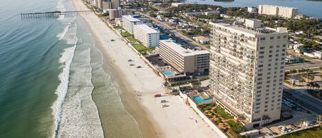 An aerial view of a coastal hotel with a beachside location, featuring multiple high-rise buildings, a pier extending into the ocean, and a clear view of the adjacent water body.