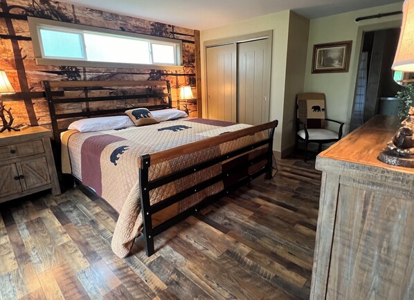 Un dormitorio con cabecera de madera, cama con colcha acolchada, mesita de noche, cómoda, silla y ventana con persianas.