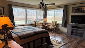 Un dormitorio con una cama grande, ventilador de techo, chimenea, televisor y vistas al exterior.