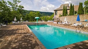 Piscina con sillas de descanso y sombrillas, rodeada por un patio de ladrillo y un muro de piedra.