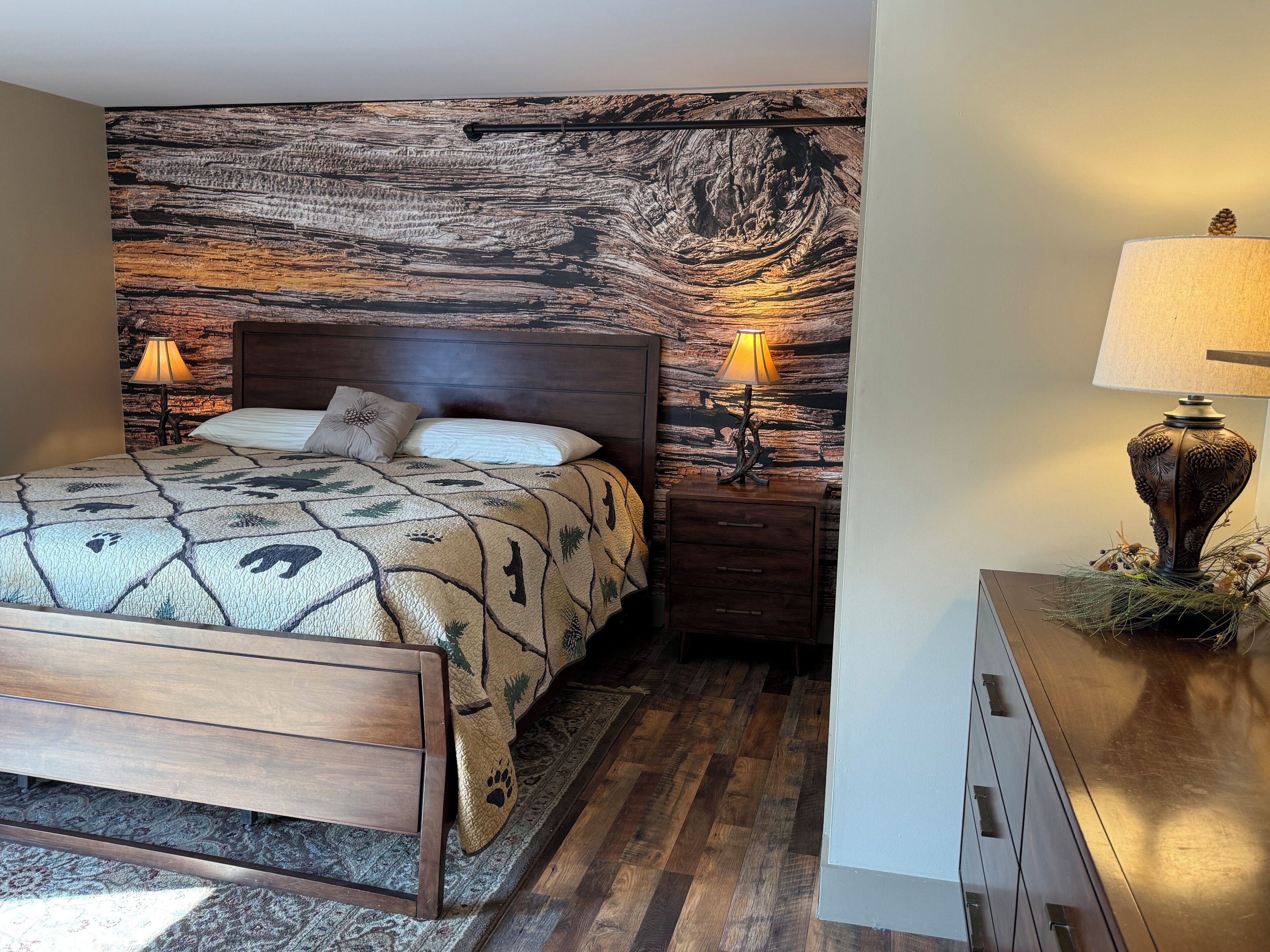 A bedroom with a wooden headboard, a patterned bedspread, two bedside lamps, and a nightstand with a decorative lamp.