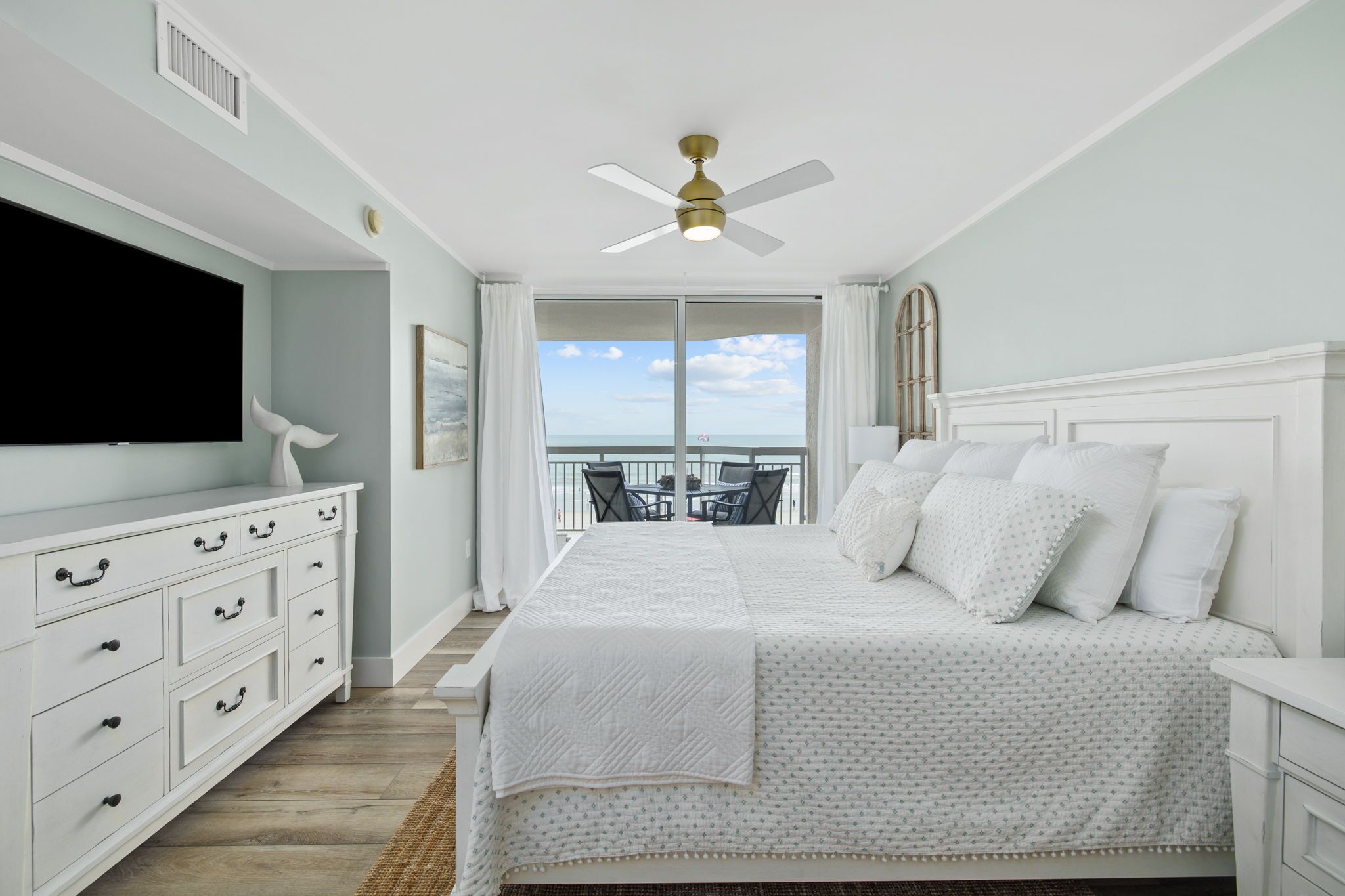 Une chambre à coucher comprenant un lit, une télévision, un ventilateur de plafond, un balcon avec vue et une commode blanche.