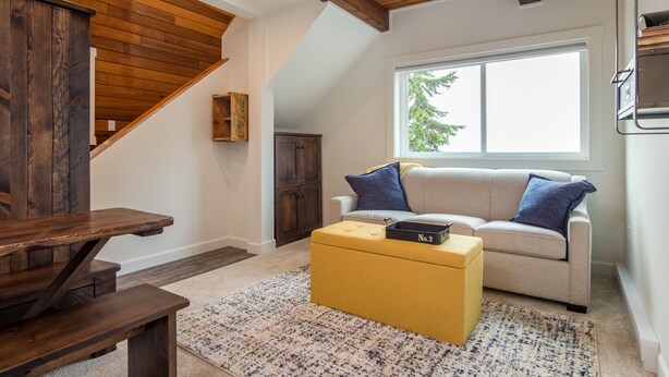 A modern living room with a wooden staircase, a sofa with blue cushions, and a yellow ottoman. - Dicky Bird Mount Washington Village (courtenay)