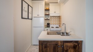 A compact kitchen with white cabinets, a white countertop, and a wooden cabinet with a metal tray. - Dicky Bird Mount Washington Village (courtenay)