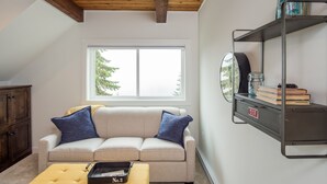 A cozy living room with a beige sofa, two blue pillows, a yellow ottoman, and a metal shelf with books and jars. - Dicky Bird Mount Washington Village (courtenay)