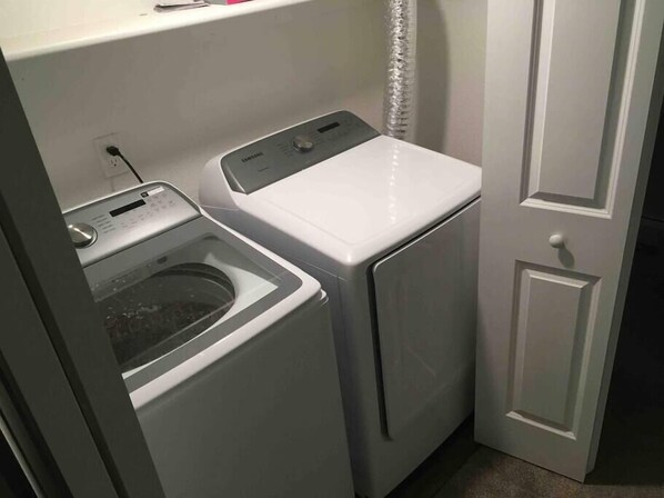 A compact laundry area with a front-loading washer and dryer, a power outlet, and a door to the right.