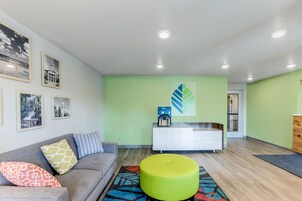 Una sala de estar moderna con un sofá gris, cojines coloridos, un reposapiés verde y una zona de cocina con un armario blanco y una cafetera.