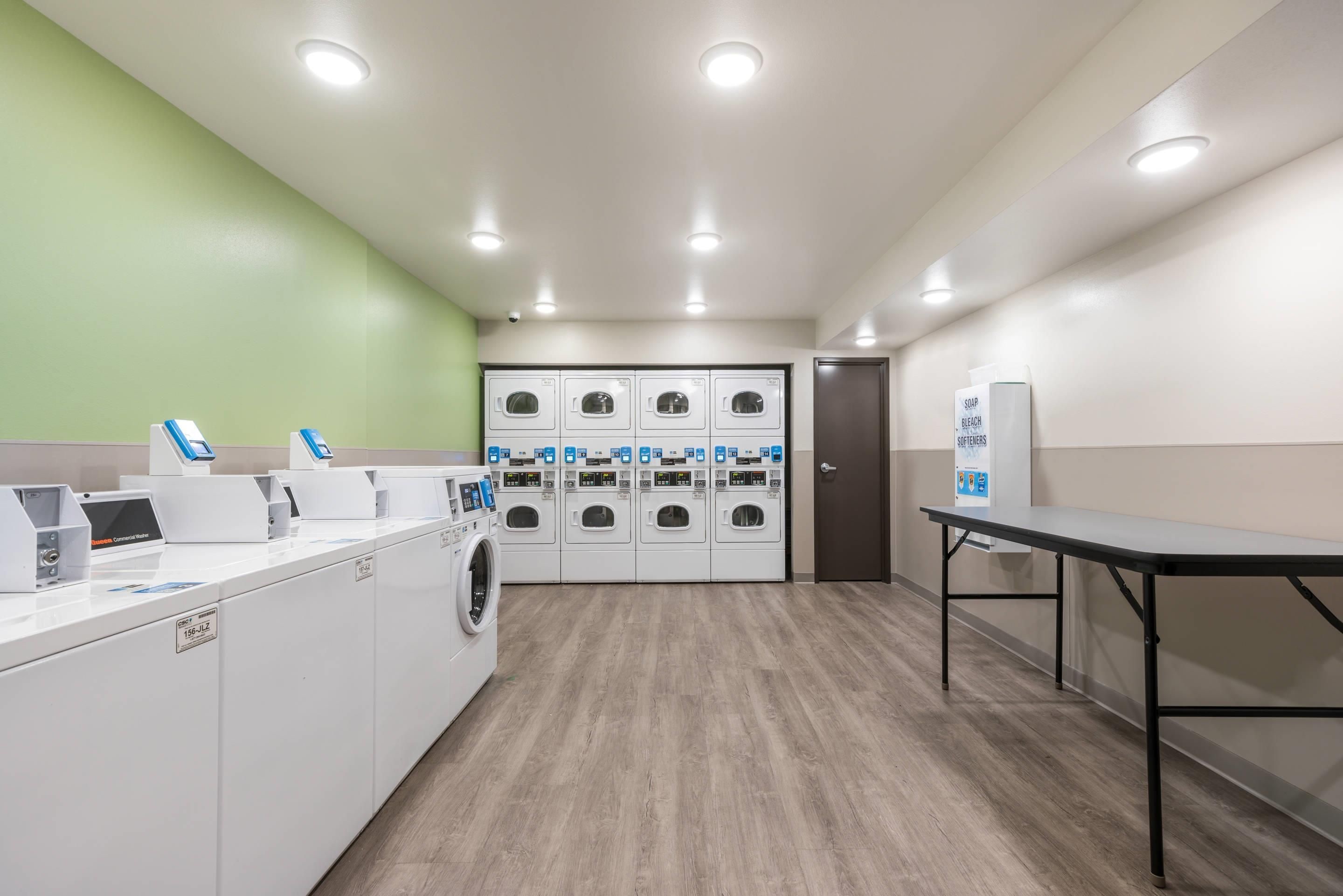 Une buanderie équipée de plusieurs machines à laver et de plusieurs s èche-linge, d’une table et d’un distributeur de savon fixé au mur.