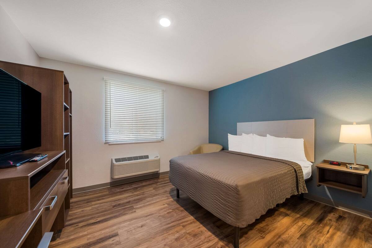 Une chambre d’hôtel avec un lit, une télévision, une table de chevet avec une lampe, une fenêtre avec des stores et un plancher en bois.
