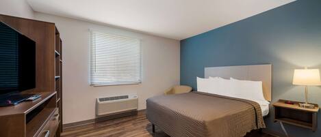 A hotel room with a bed, a TV, a bedside table with a lamp, a window with blinds, and a wooden floor.