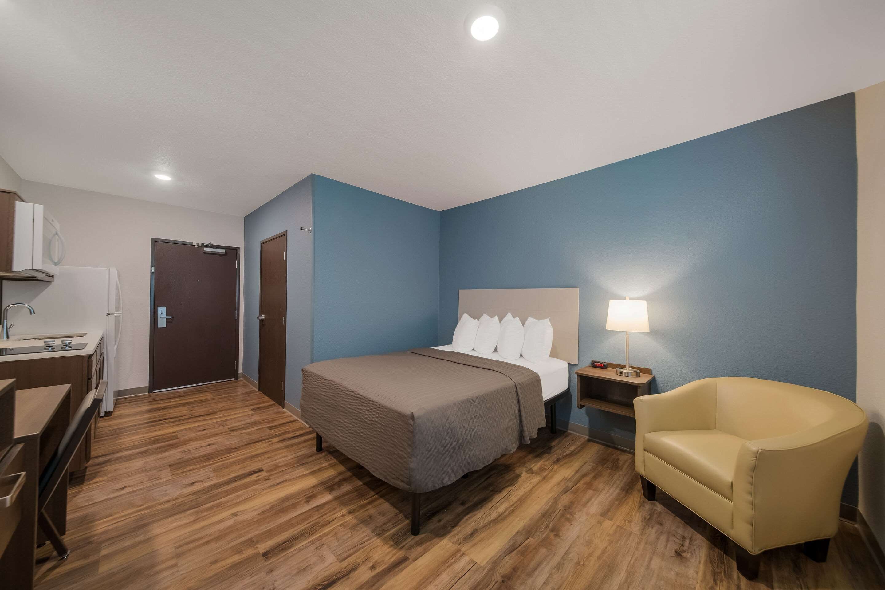 A hotel room with a bed, bedside table, lamp, chair, and a desk with a computer.