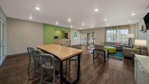 A modern living room with a wooden dining table, grey chairs, a grey sofa, and a green ottoman. - WoodSpring Suites Wesley Chapel-Tampa (Wesley Chapel)