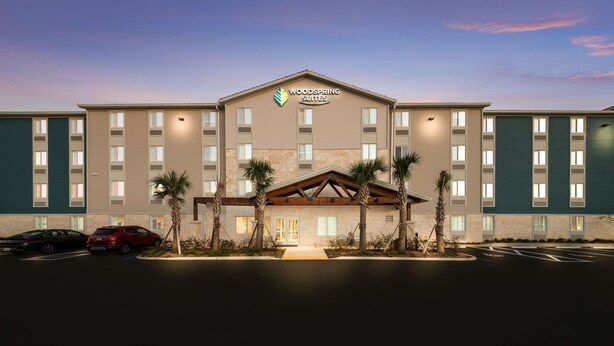 A multi-story hotel with a prominent logo, a central entrance, and a parking lot with several cars. - WoodSpring Suites Wesley Chapel-Tampa (Wesley Chapel)
