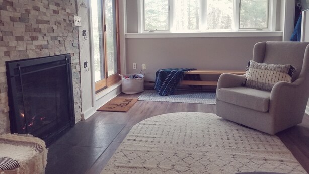Ein Wohnzimmer mit Kamin, einem bequemen Sessel, einem Teppich und Blick ins Freie durch große Fenster.