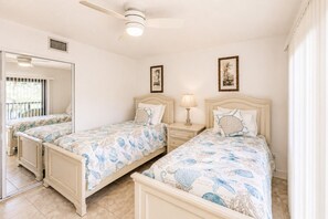 A bedroom with two beds, a ceiling fan, a nightstand with a lamp, and a window with blinds.