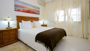 Una cama bien hecha con un edredón marrón, dos lámparas de noche, una mesita de noche de madera y un cuadro de un atardecer sobre un puente.