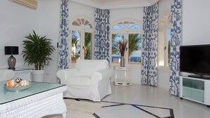 Una sala de estar con un sillón blanco, una mesa de centro de vidrio con fruta, un televisor y vista al mar a través de cortinas estampadas.