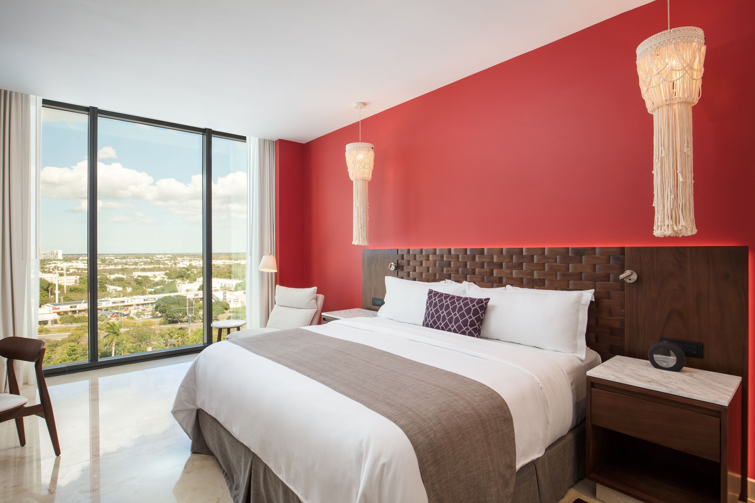 Une chambre d’hôtel moderne dotée d’un grand lit, d’une tête de lit en bois et offrant une vue sur la ville grâce à de grandes fenêtres du sol au plafond.