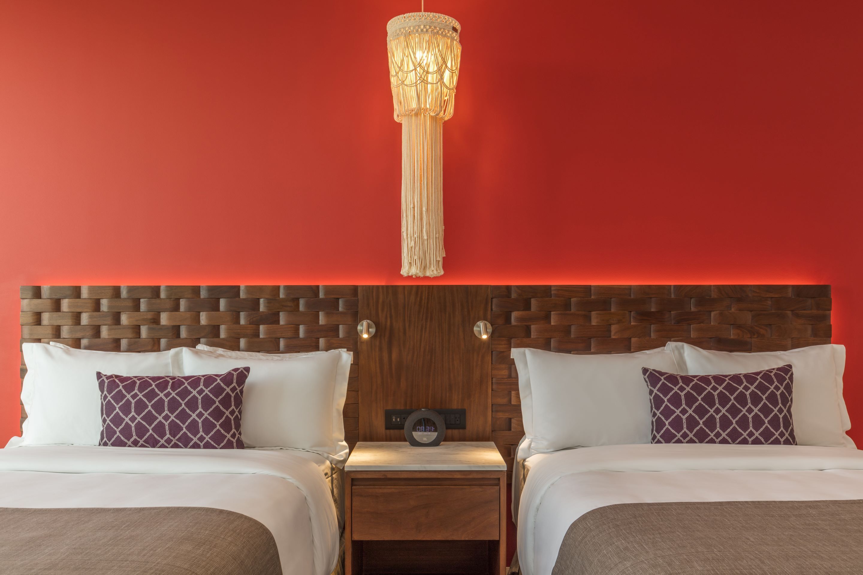 Une chambre d’hôtel avec une tête de lit en bois, deux lits avec du linge de lit blanc et des oreillers à motifs violets, une table de chevet avec une horloge et un luminaire suspendu.