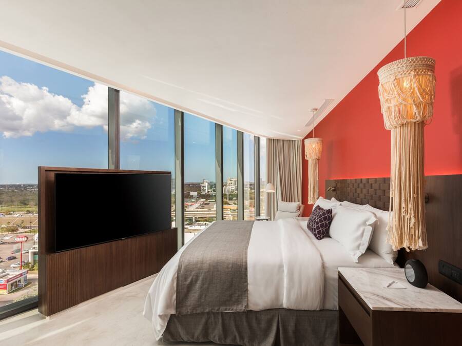 A modern hotel room with a large bed, a flat-screen TV, and a city view through large windows.