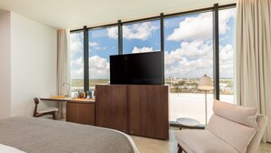 A hotel room with a large window, a bed, a desk with a television, and a sofa. - Camino Real Mérida (Mérida)