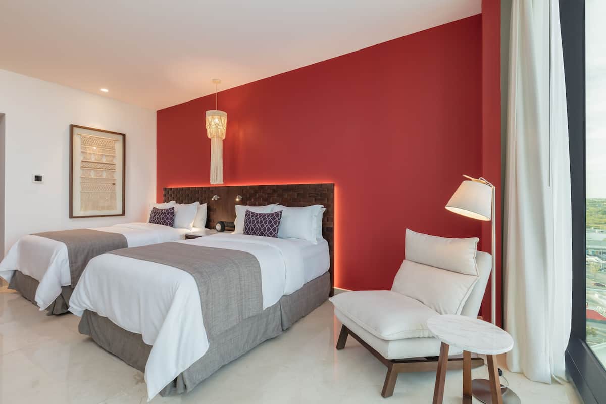 A hotel room with two beds, a red accent wall, a modern chandelier, a white armchair, and a view of the city.