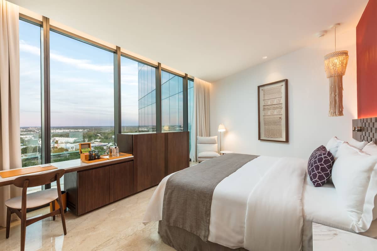 A modern hotel room with a large window, a desk with a chair, a bed with pillows, and a hanging light fixture.