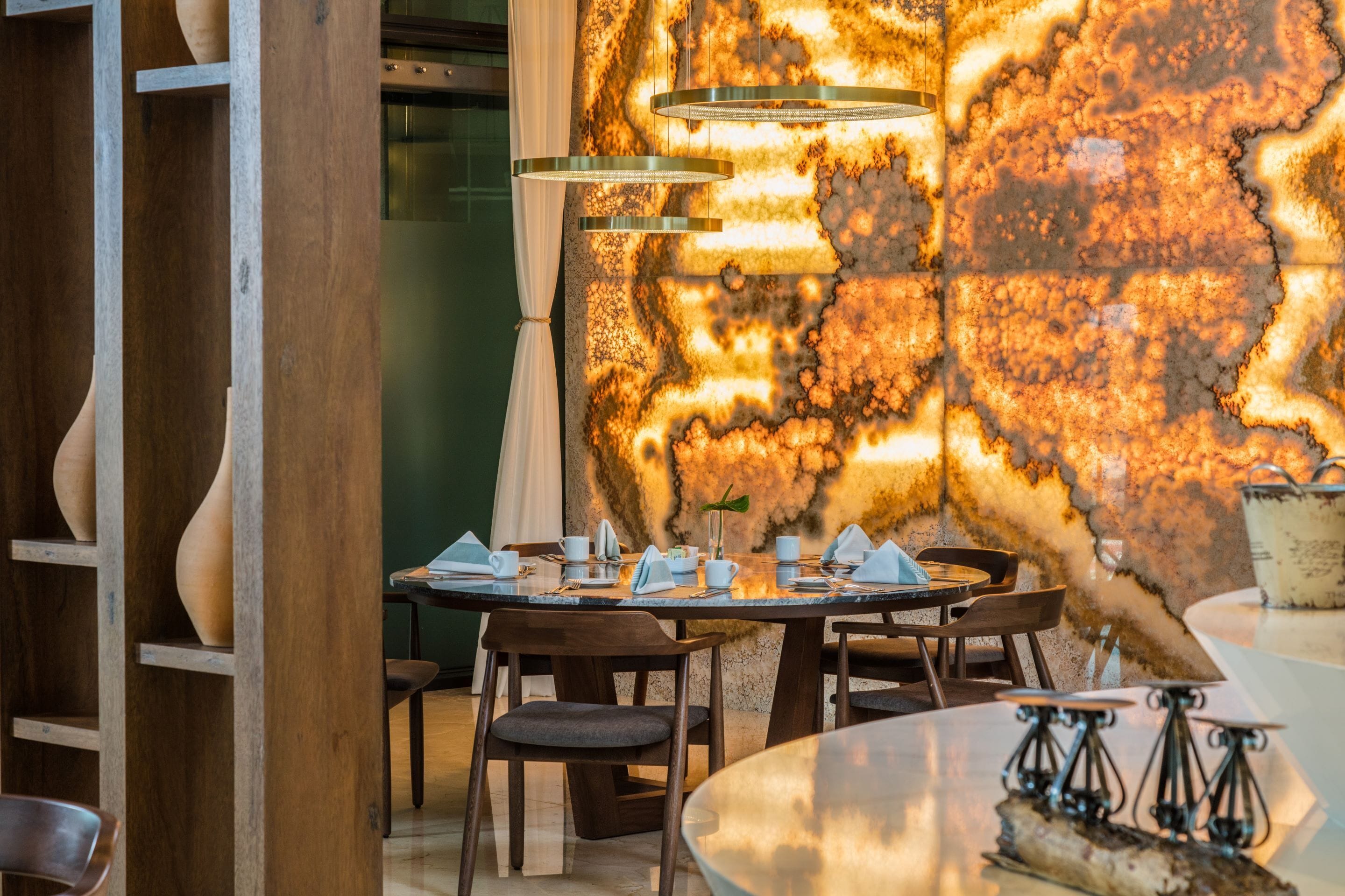 A restaurant interior with a round table set for dining, wooden chairs, and a decorative wall with a textured pattern.