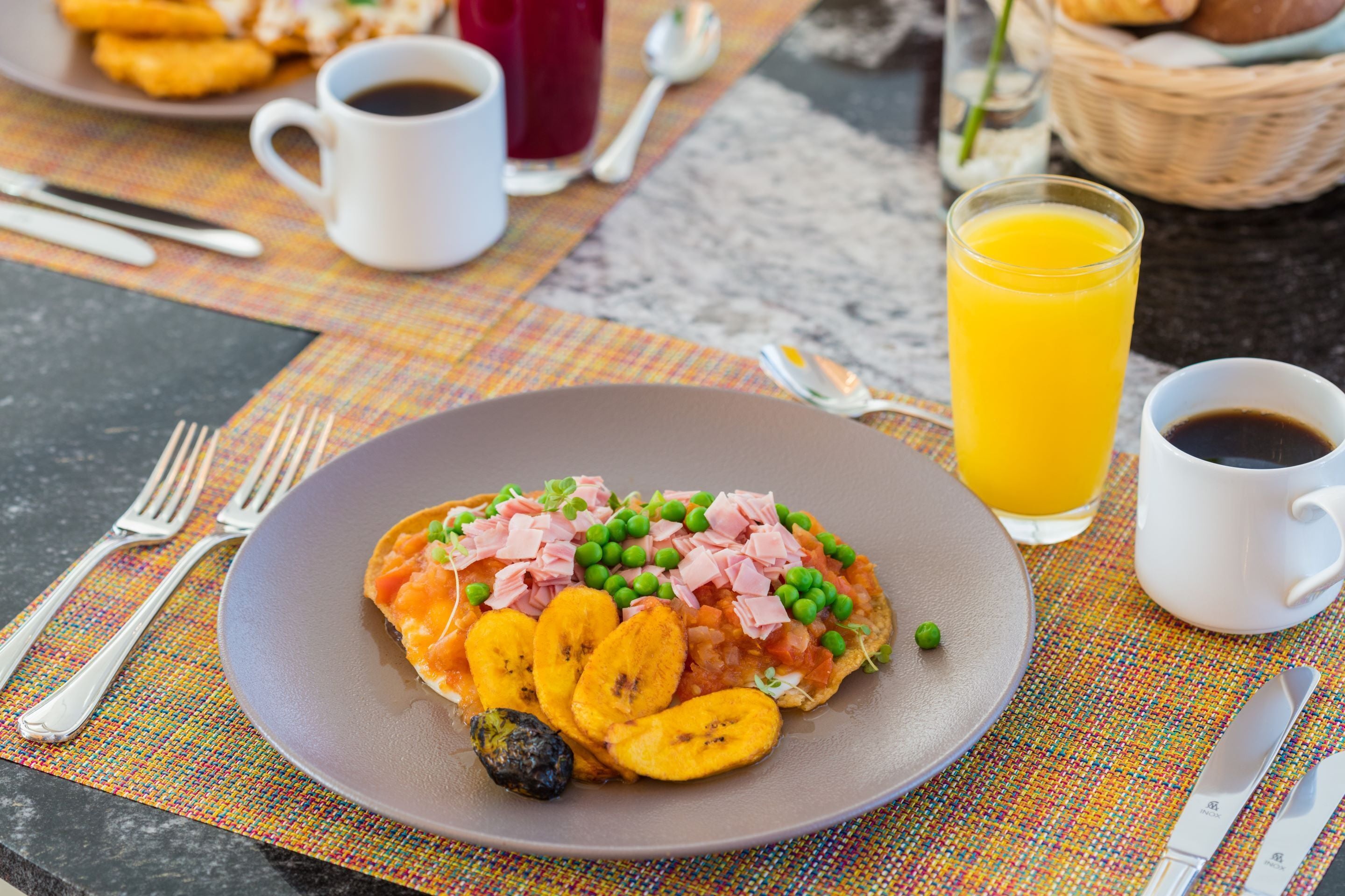 A breakfast plate with ham, peas, and bananas, accompanied by a glass of orange juice and two cups of coffee on a table with a colorful placemat.