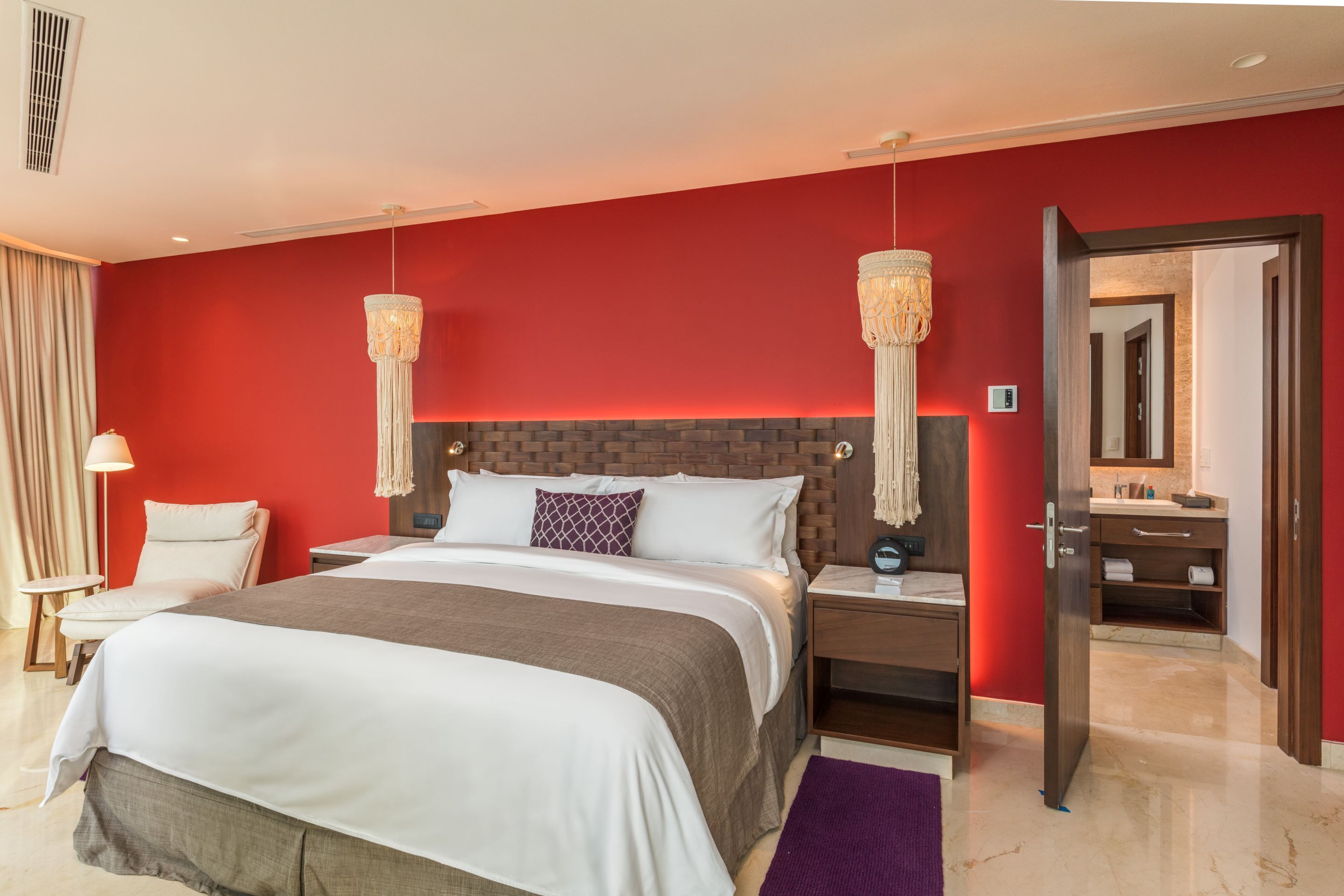 A modern hotel room with a large bed, a wooden headboard, two bedside lamps, a nightstand, a bathroom visible through an open door, and a red accent wall.
