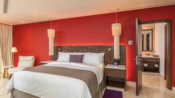 A modern hotel room with a large bed, a wooden headboard, two bedside lamps, a nightstand, a bathroom visible through an open door, and a red accent wall.