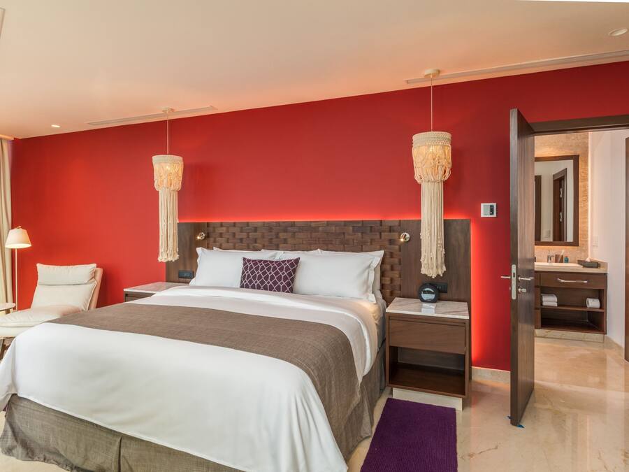 A modern hotel room with a large bed, a wooden headboard, two bedside lamps, a nightstand, a bathroom visible through an open door, and a red accent wall.