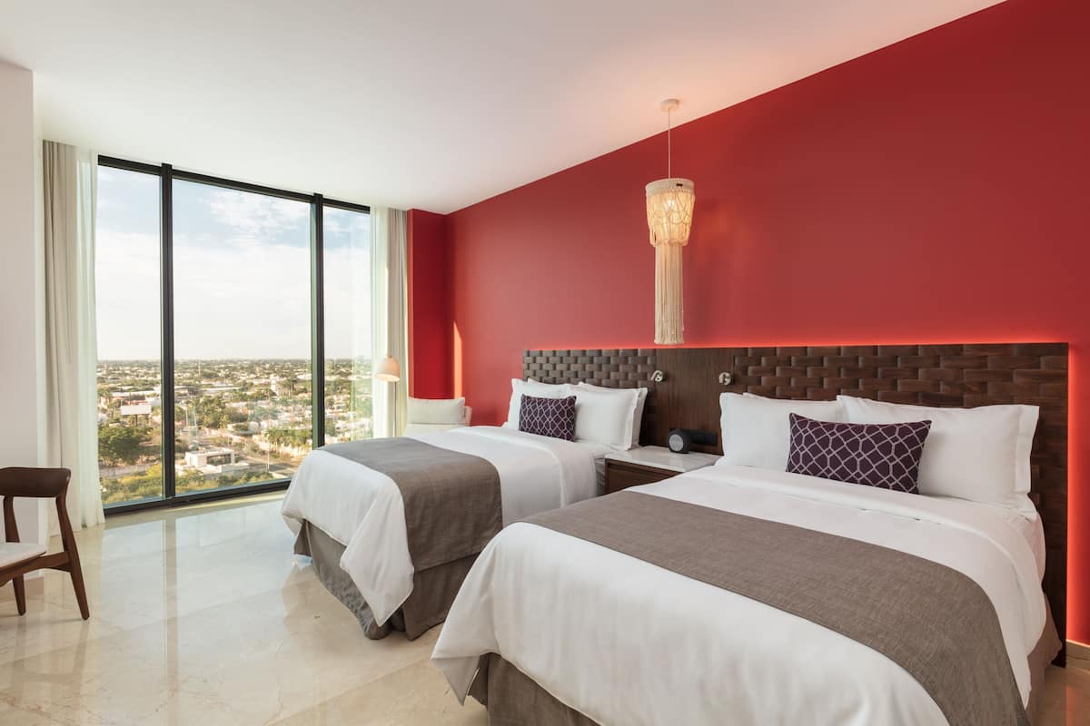 A hotel room with two beds, a large window with a city view, a red accent wall, and a modern chandelier.