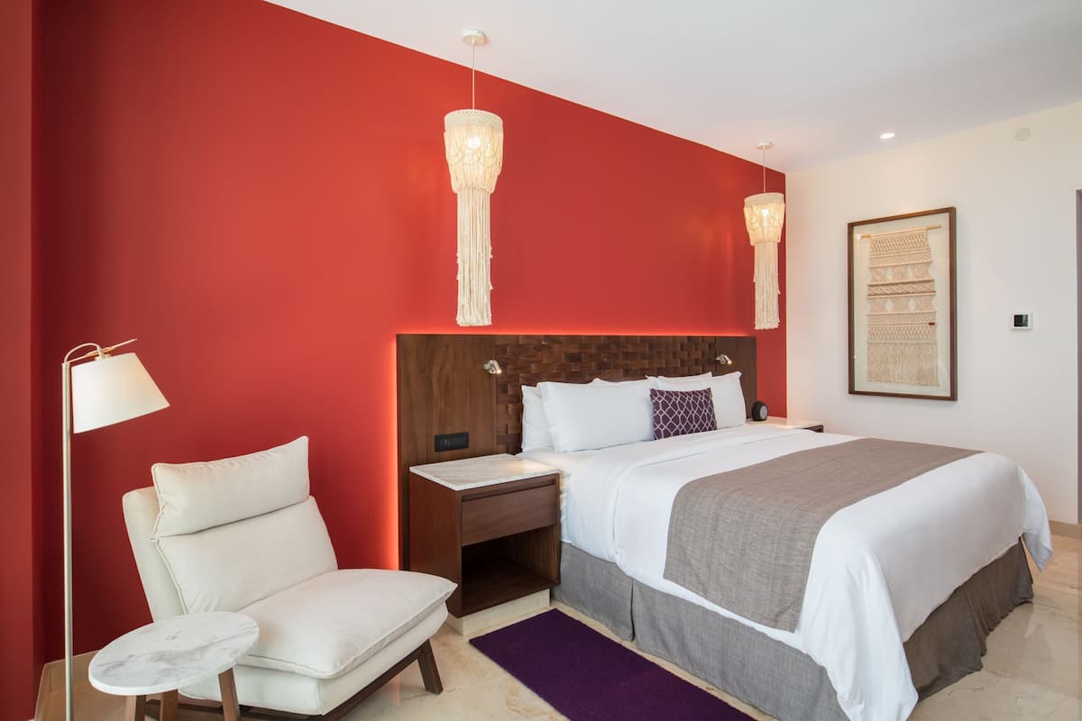 A modern bedroom with a red accent wall, a large bed with white bedding, a nightstand, a chair, and two pendant lights.