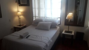 Una cama bien hecha con sábanas blancas, dos lámparas de noche y un espejo grande en la pared.