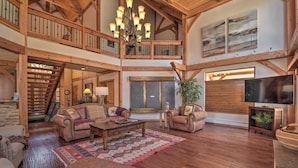 A spacious living room with a high ceiling, wooden beams, a chandelier, leather sofas, a flat-screen TV, and a staircase leading to an upper level. - Grand Angel Fire Retreat w/ Hot Tub & Fire Pit! (Angel Fire)
