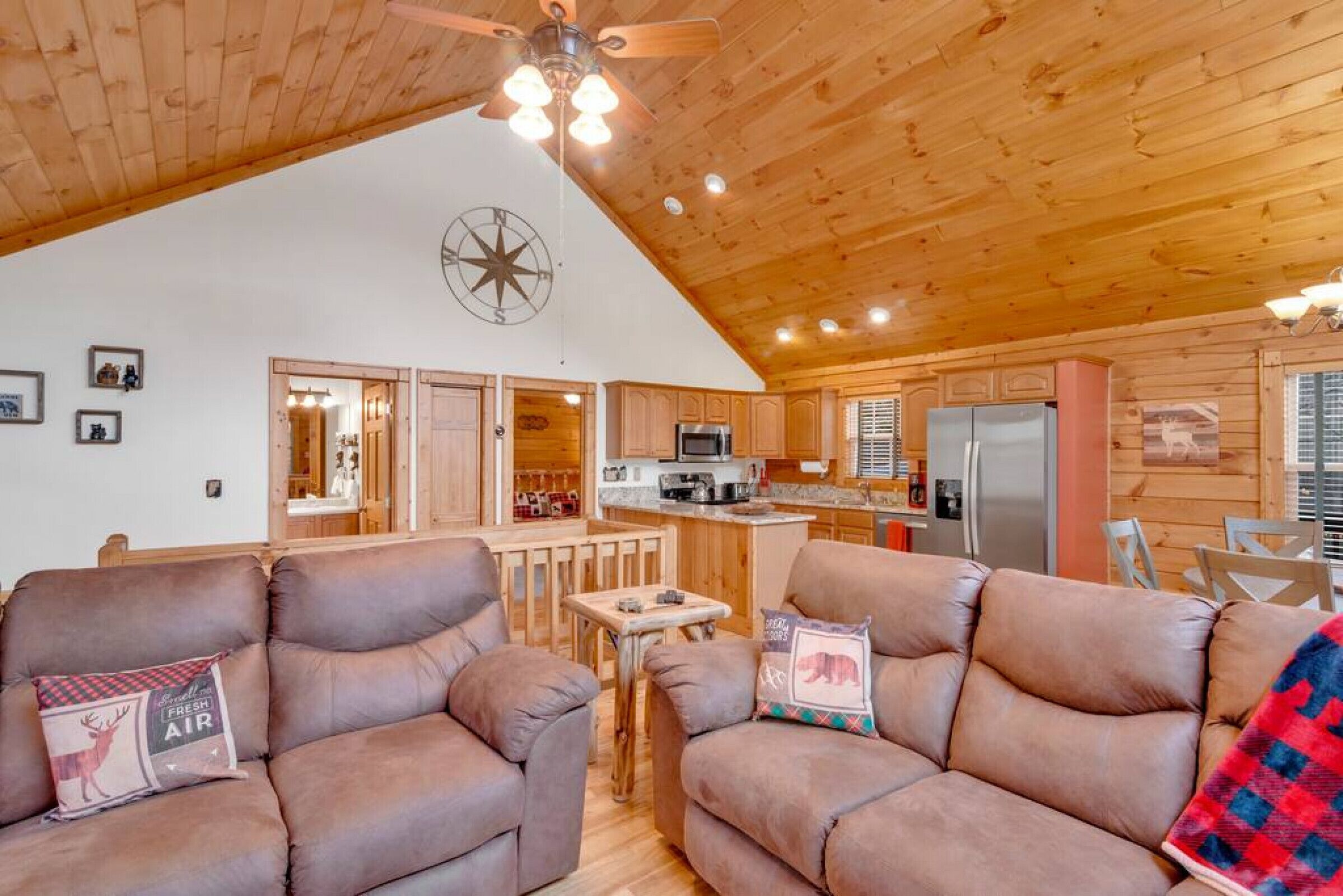 L’intérieur chaleureux d’un chalet avec un plafond haut, des murs en bois, un coin cuisine et deux canapés en cuir agrémentés de coussins décoratifs.