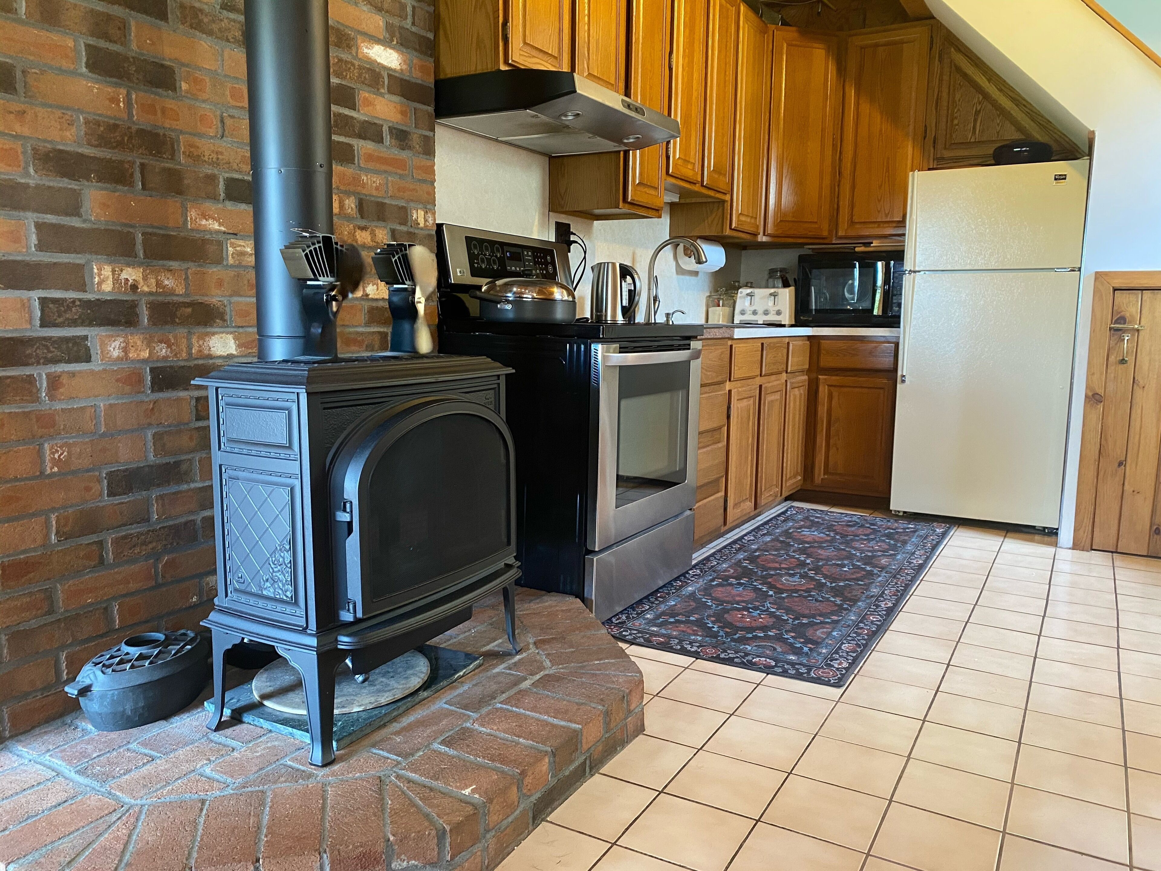 A kitchen with a brick wall, a wood stove, a stainless steel oven, wooden cabinets, and a tiled floor.
