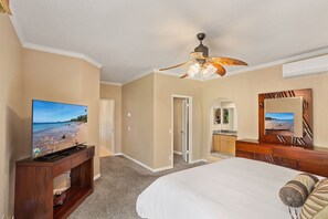 A hotel room with a large bed, a ceiling fan, a TV on a wooden console, a bathroom with a mirror, and a view of the beach through the door.