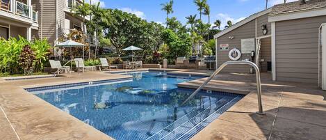 A swimming pool with lounge chairs and umbrellas, surrounded by palm trees and a building with balconies.