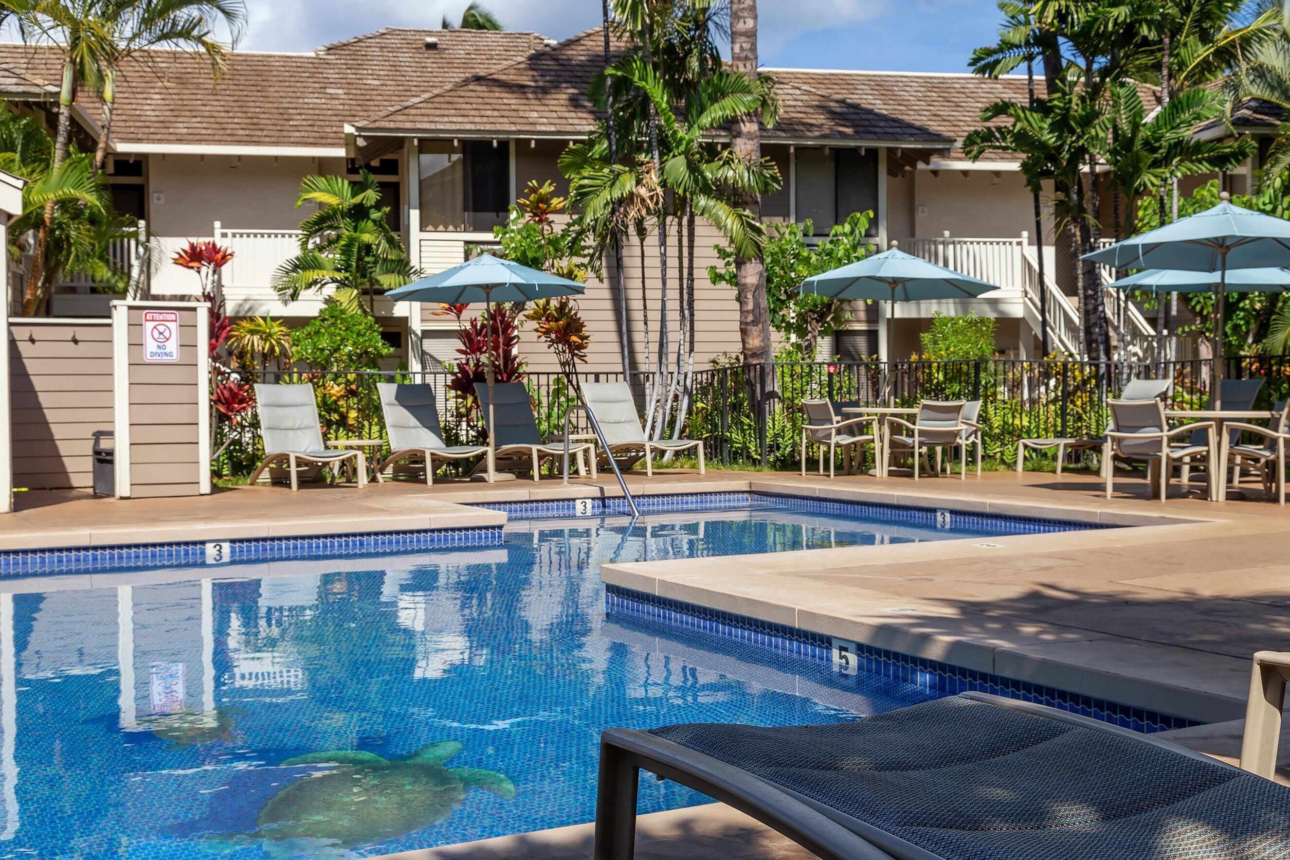 A swimming pool with lounge chairs and umbrellas, surrounded by palm trees and a building with a tiled roof.